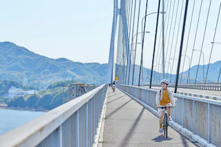 A person wearing a helmet rides a bicycle on a bridge with mountains and water visible in the background; other cyclists are seen further along the bridge under a clear blue sky.