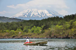 A person wearing a hat rows a small boat on a calm lake, surrounded by green trees, with snow-capped Mount Fuji towering in the background under a partly cloudy sky.