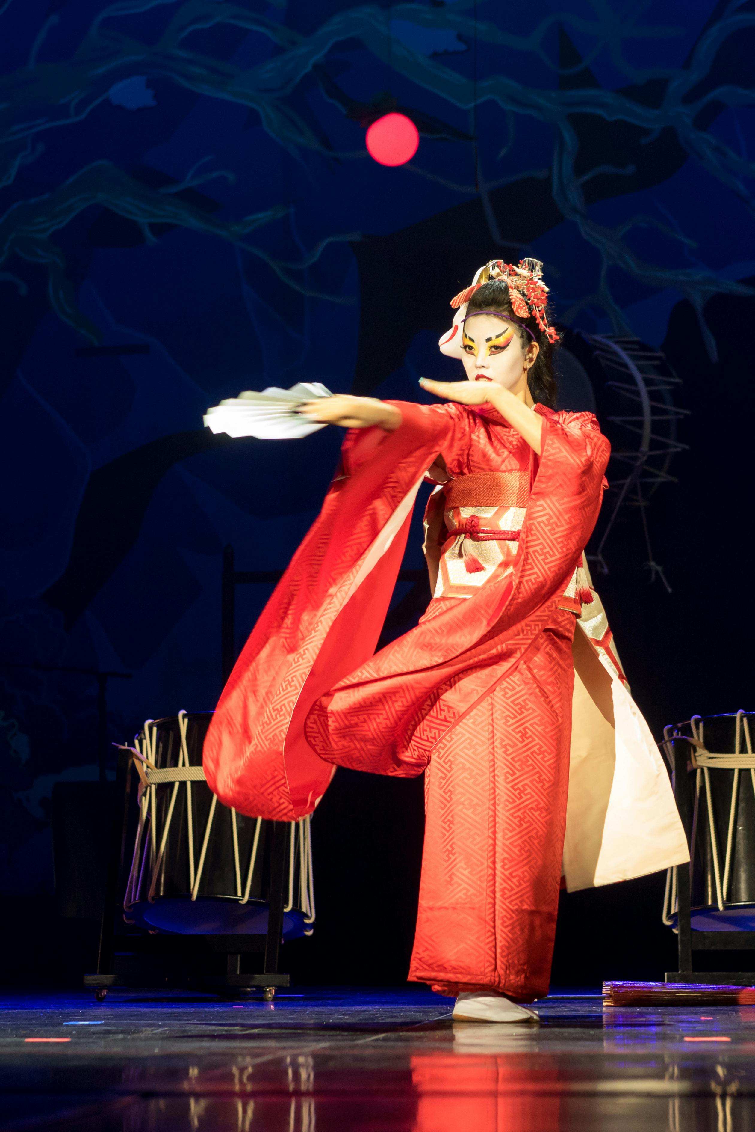 A performer in a red kimono and traditional makeup dances on stage, holding a white fan, with drums and a moonlit backdrop behind them.