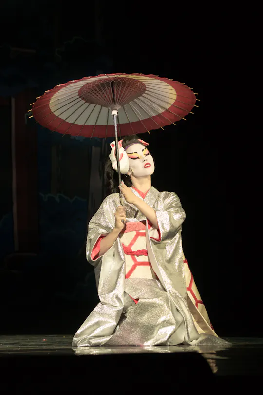 A performer in a silver kimono and traditional white face makeup kneels on stage, holding a red and white paper umbrella above her shoulder. The background is dark, emphasizing the dramatic lighting on her.