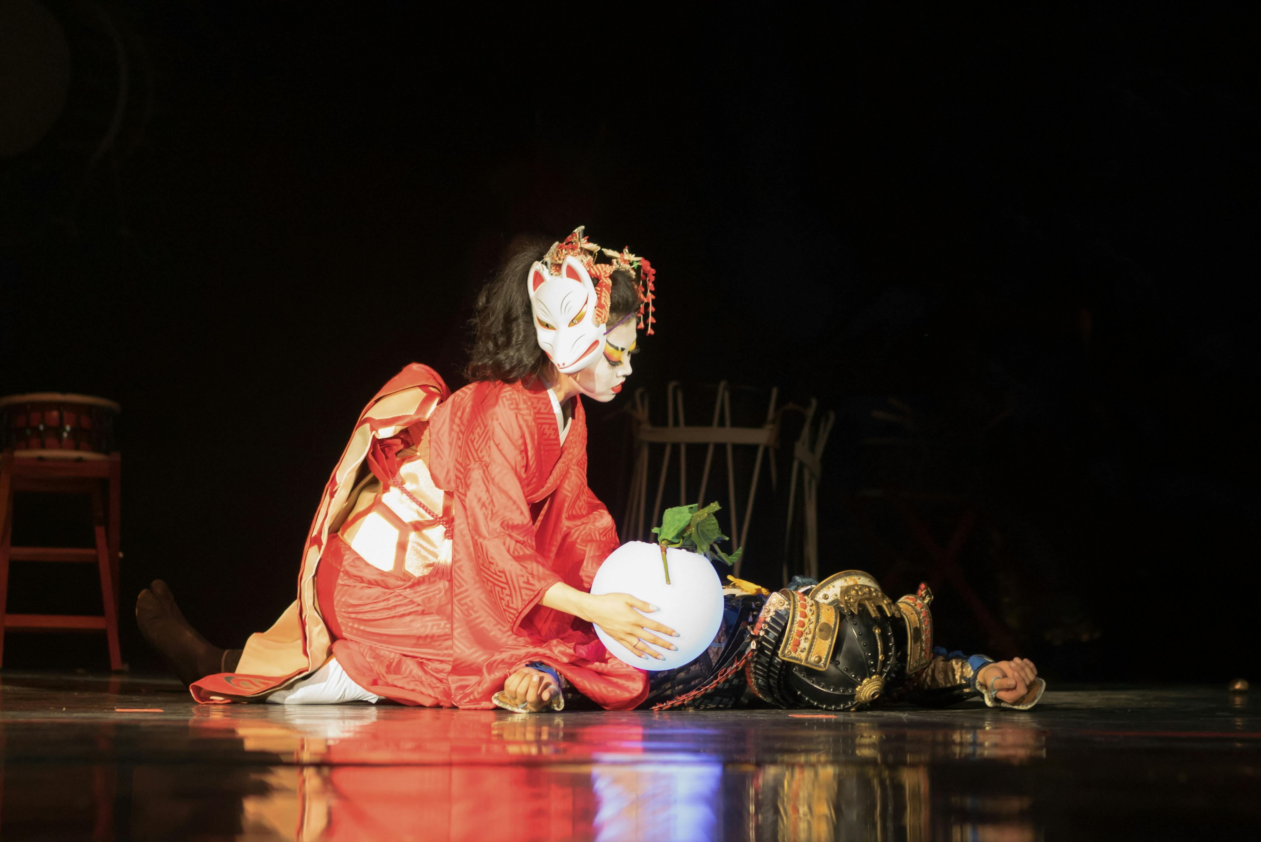 A performer in a red kimono and fox mask kneels beside a person in samurai armor lying on stage, holding a glowing white sphere with a green leaf. The scene is dramatically lit against a dark background.