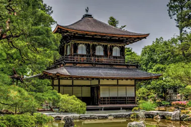 A traditional Japanese wooden pavilion with a dark tiled roof, surrounded by lush green trees and a tranquil pond in the foreground, under a cloudy sky.