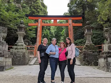 Four women stand together posing in front of a large red torii gate surrounded by lush greenery and stone lanterns at a traditional Japanese shrine.