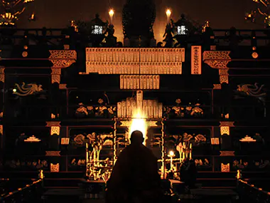 A person sits in front of an ornate altar illuminated by candlelight, with golden decorations, intricate carvings, and tablets, creating a warm, spiritual atmosphere.