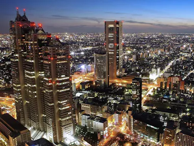 Aerial view of a brightly lit city at dusk, featuring tall skyscrapers with red lights on top, illuminated streets, and buildings stretching into the horizon under a blue evening sky.