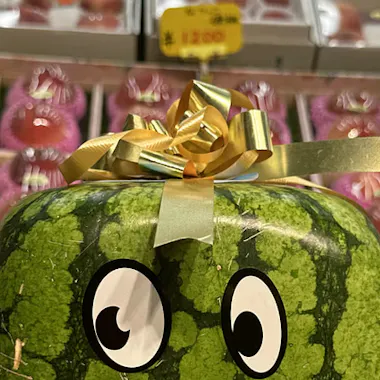 Tokyo Tour A square watermelon with cartoon eyes and a gold ribbon on top is displayed in a market, with neatly arranged fruit packages and a yellow price sign in the background.