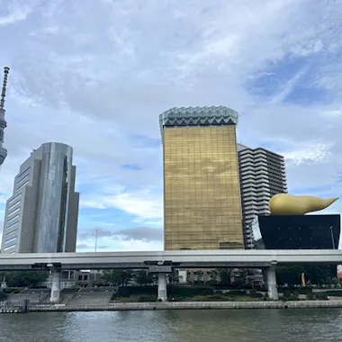 Tokyo Tour A riverside cityscape featuring the Tokyo Skytree, modern buildings, and the Asahi Beer Hall with its distinctive golden flame sculpture under a partly cloudy sky.