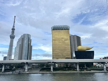 A riverside cityscape featuring the Tokyo Skytree, modern buildings, and the Asahi Beer Hall with its distinctive golden flame sculpture under a partly cloudy sky.