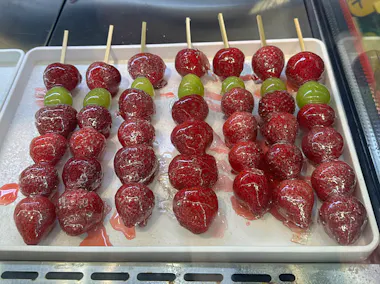 Several skewers of candied strawberries and green grapes are arranged neatly on a white tray. The fruits are coated in a shiny, clear sugar glaze.