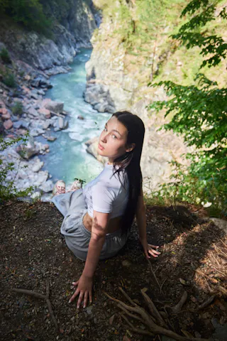 A woman with long dark hair sits on a rocky ledge overlooking a river winding through a forested canyon, sunlight illuminating her face and casting shadows on the ground.