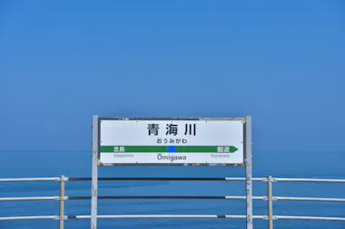 A white sign for Ōmigawa Station in Japanese and English stands on a seaside platform with blue sky and calm ocean in the background. Directional signs for Kasaishiwa and Kujiranami are also visible.