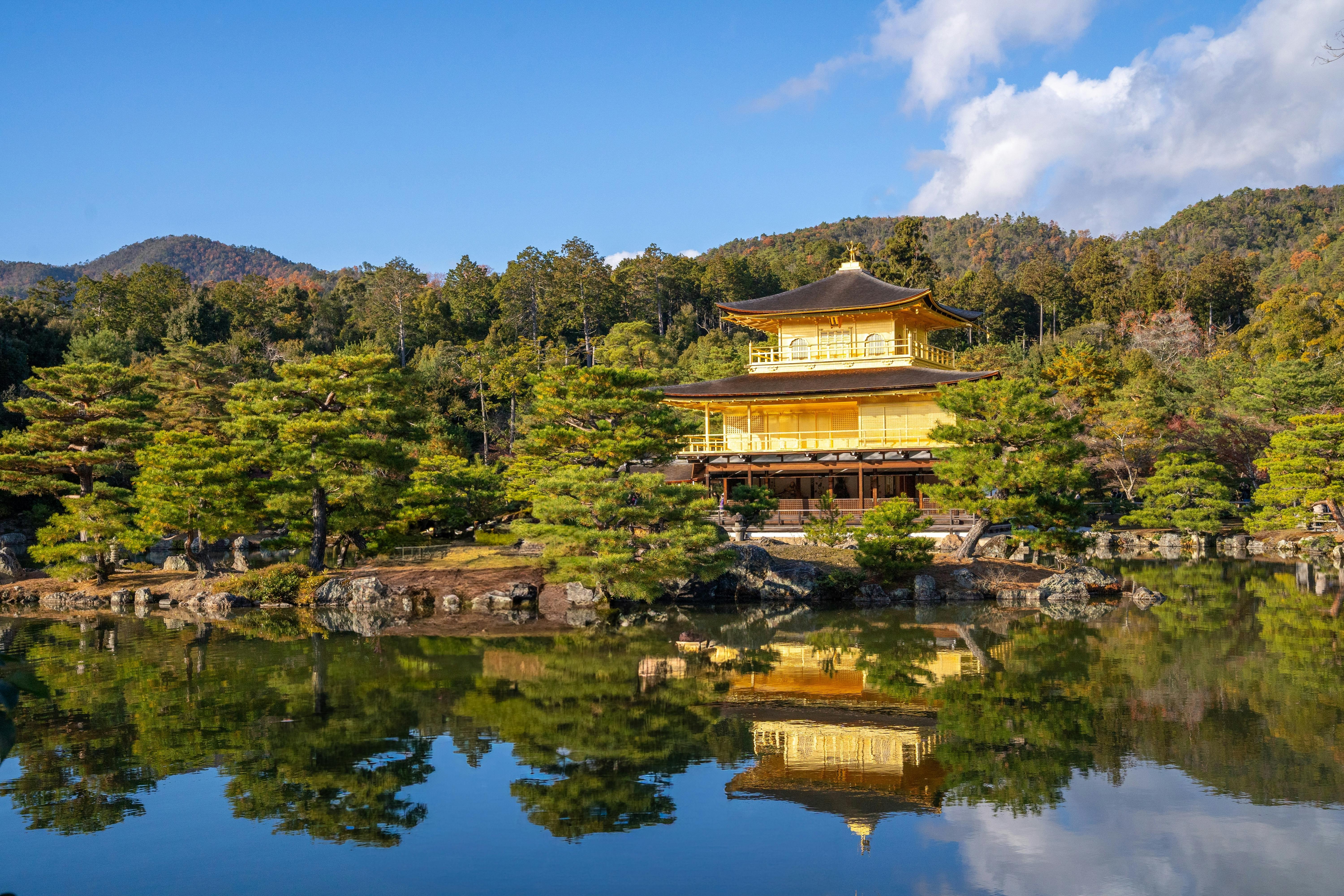 A golden pavilion surrounded by trees is reflected in a calm pond under a blue sky, with forested hills in the background.