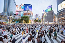 A bustling city scene at a large intersection with numerous pedestrians crossing. Surrounding buildings display bright advertisements and electronic billboards. The sky is overcast, and the atmosphere is vibrant and lively.