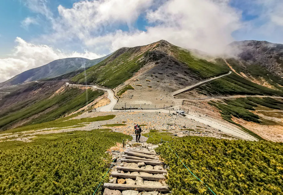 一个人站在一条通往崎岖山地的木栈道上，周围有零星的植被。蜿蜒的道路和云层部分遮住了在明亮蓝天下的山峰。
