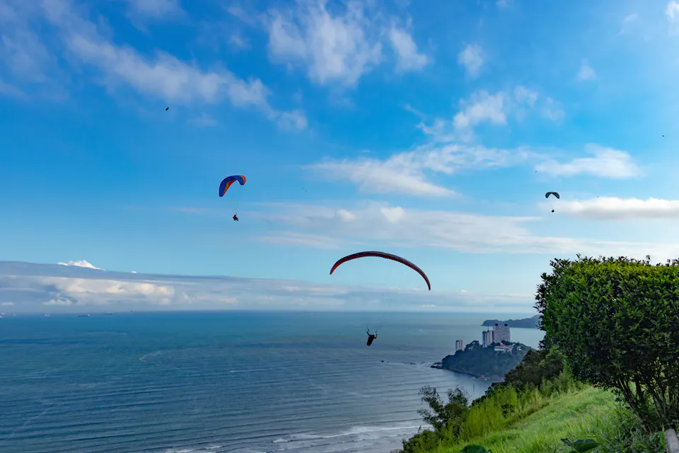 滑翔伞在海岸悬崖上空翱翔，俯瞰着海洋和远处的天际线。天空晴朗，云朵稀少，悬崖上的郁郁葱葱的绿 foliage 可见。