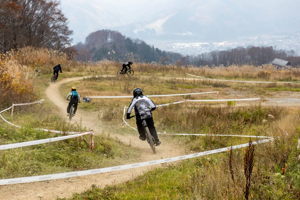 骑自行车的人穿着保护装备，在山坡上沿着弯曲的泥土小径行进。风景被秋天的树叶和遥远的山脉环绕，头上是模糊的天空。多个骑行者在小径的不同点可见。