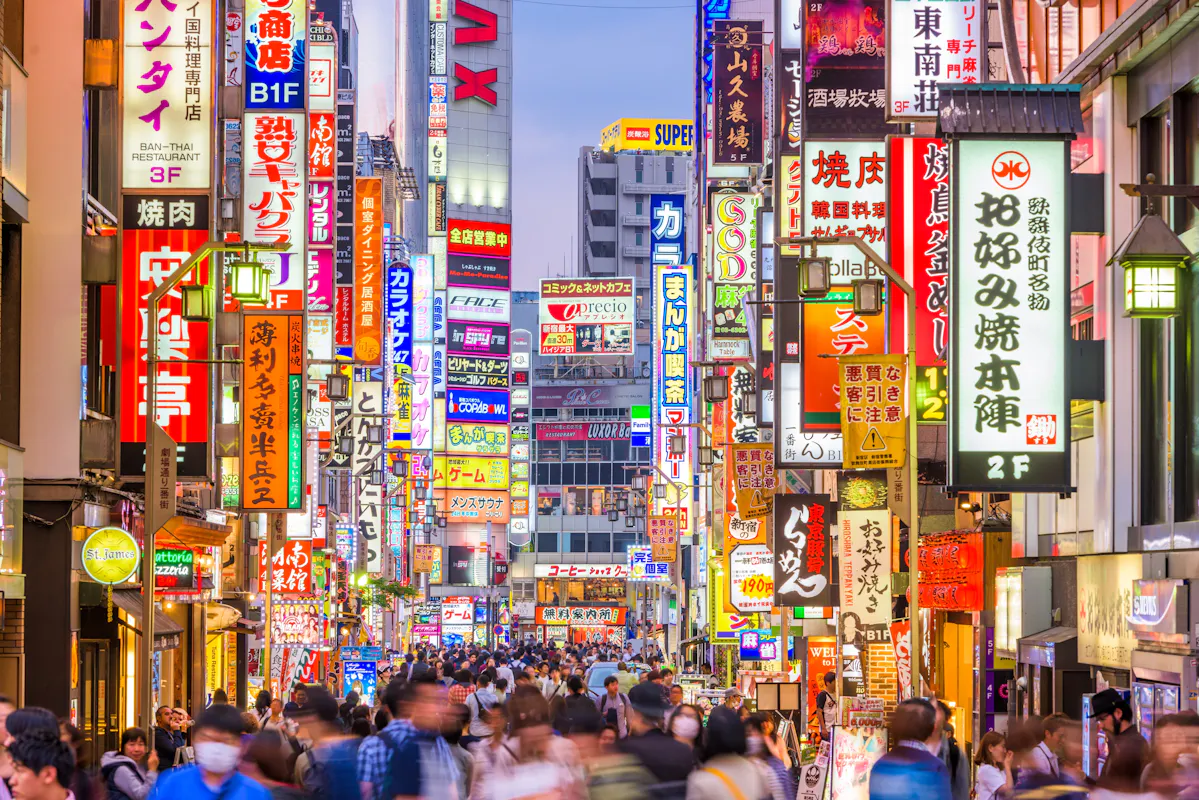 A bustling street in Tokyo, Japan, filled with people and vibrant illuminated signs in various colors. The signs are in Japanese, featuring ads for shops, restaurants, and entertainment. The vibrant atmosphere suggests a lively urban area.