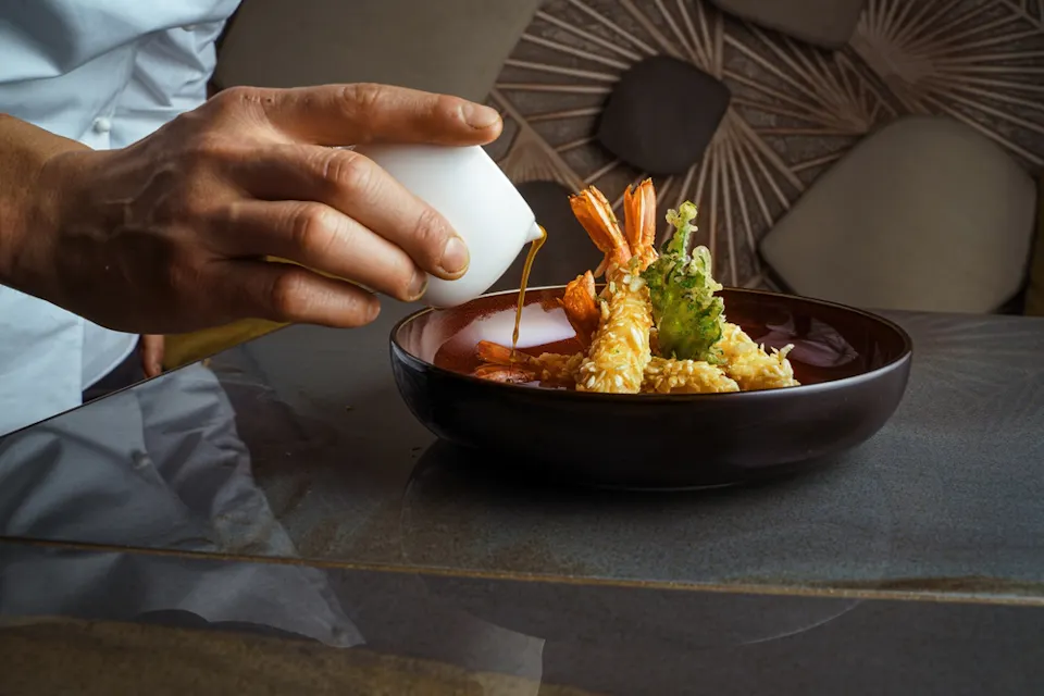 A hand pours sauce from a small white pitcher onto a dish featuring fried shrimp and tempura vegetables on a dark plate. The background shows a textured wall with abstract designs in earthy tones.