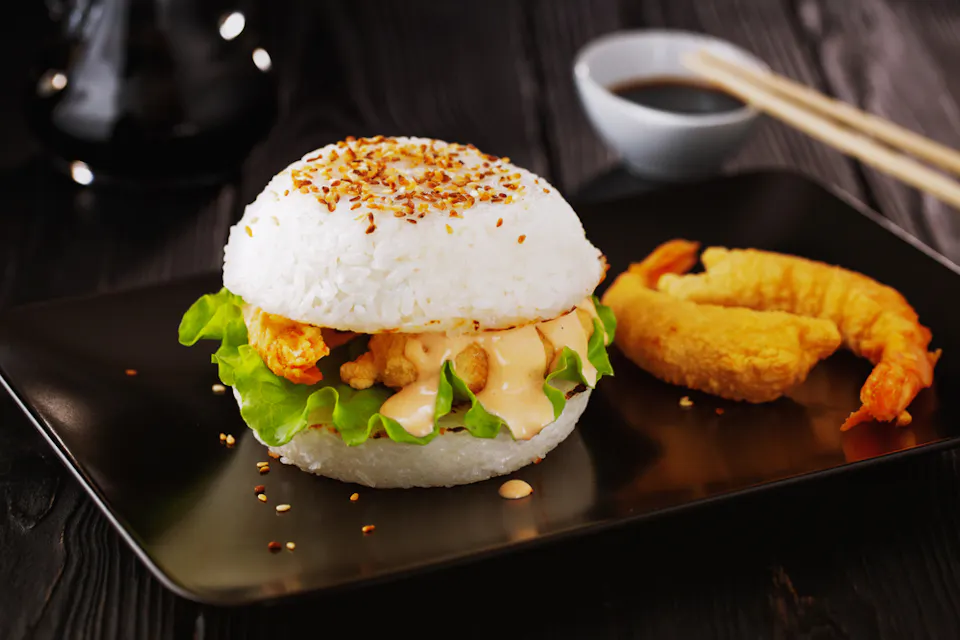A rice burger with lettuce and fried shrimp, topped with sesame seeds, on a black plate. Next to it are two pieces of tempura shrimp. A dish of soy sauce with chopsticks is in the background on a dark table.