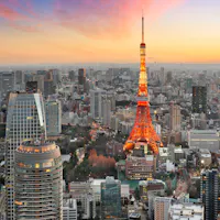 Things to Do in Tokyo Aerial view of Tokyo at sunset, featuring Tokyo Tower illuminated in orange. Surrounding skyscrapers and cityscape are bathed in warm, glowing hues, with a mix of modern architecture and lush greenery. A pink and orange sky adds to the scene's vibrancy.