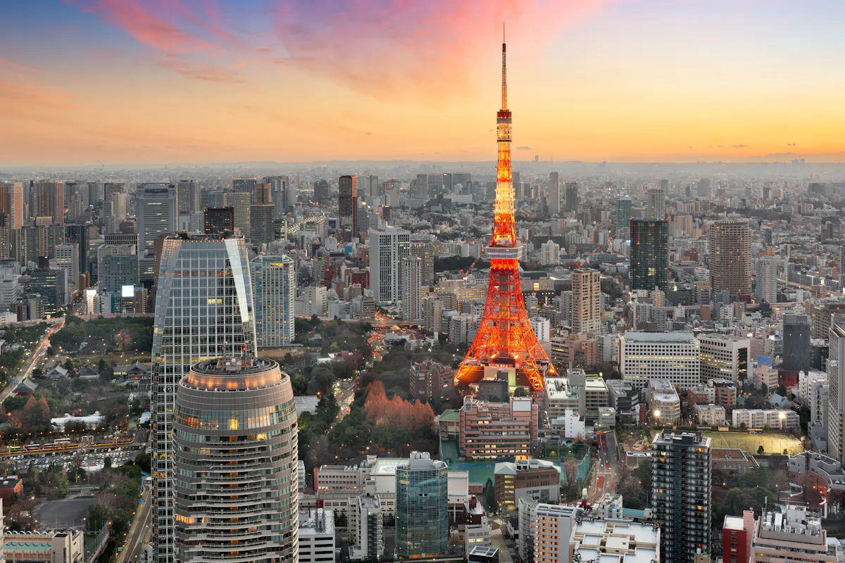 Things to Do in Tokyo Aerial view of Tokyo at sunset, featuring Tokyo Tower illuminated in orange. Surrounding skyscrapers and cityscape are bathed in warm, glowing hues, with a mix of modern architecture and lush greenery. A pink and orange sky adds to the scene's vibrancy.