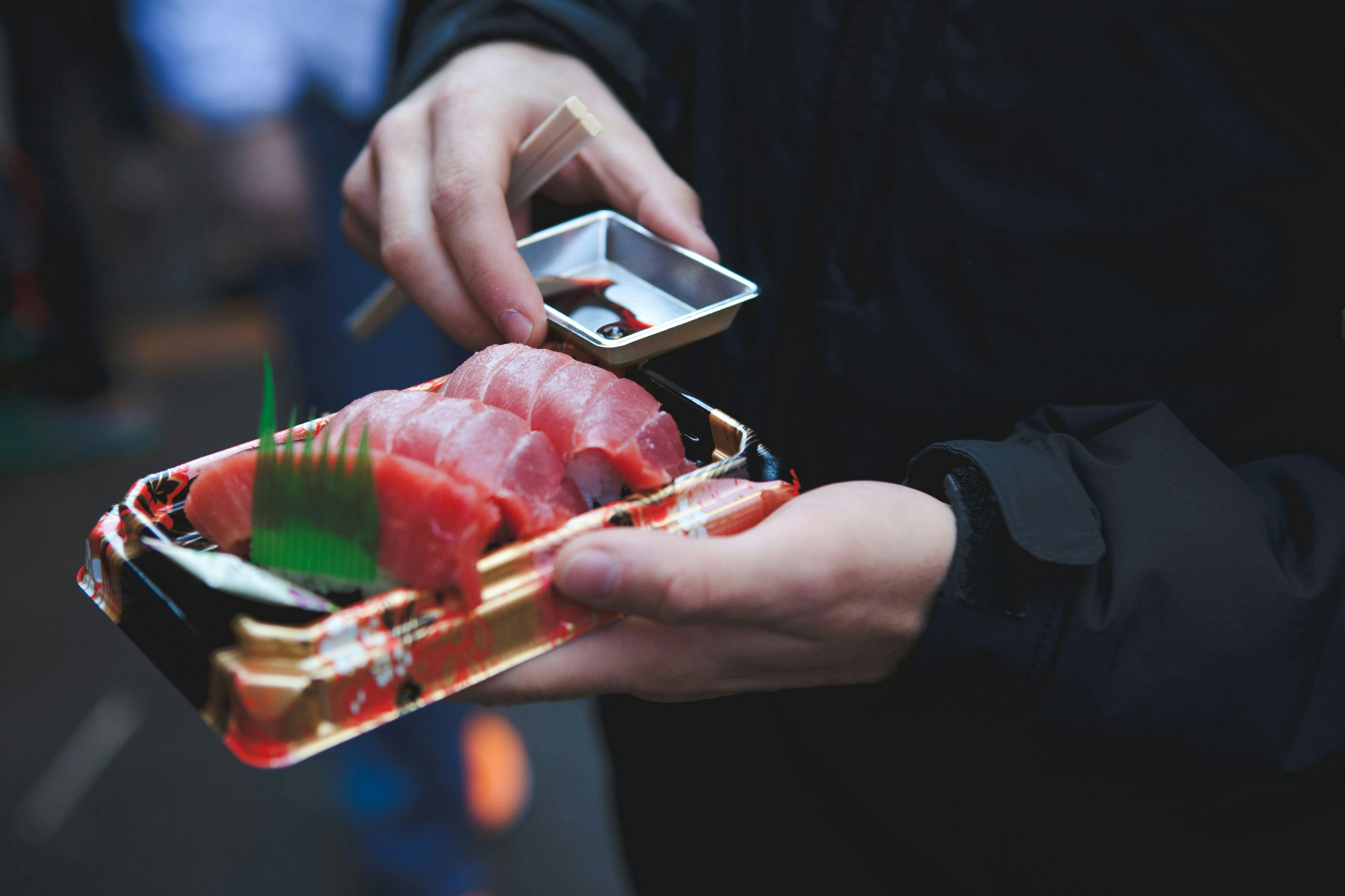 A person holding a tray of sliced raw fish, likely sashimi, in one hand and dipping sauce in the other, with chopsticks resting on the sauce container.