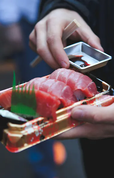 Tsukiji Fish Market A person holding a tray of sliced raw fish, likely sashimi, in one hand and dipping sauce in the other, with chopsticks resting on the sauce container.