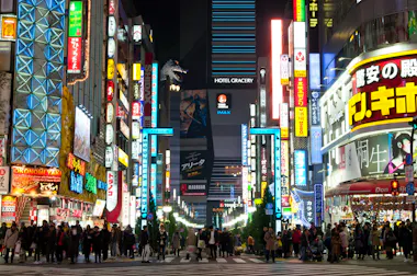 A busy street in Tokyo at night, filled with crowds of people and lined with bright, colorful neon signs in Japanese. A large Godzilla figure is visible on a building in the background above the “Hotel Gracery” sign.