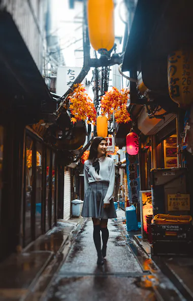 Shinjuku Golden Gai A woman walks down a narrow, lantern-lit alley lined with small shops and restaurants, with colorful signs and hanging decorations, on a rainy day.