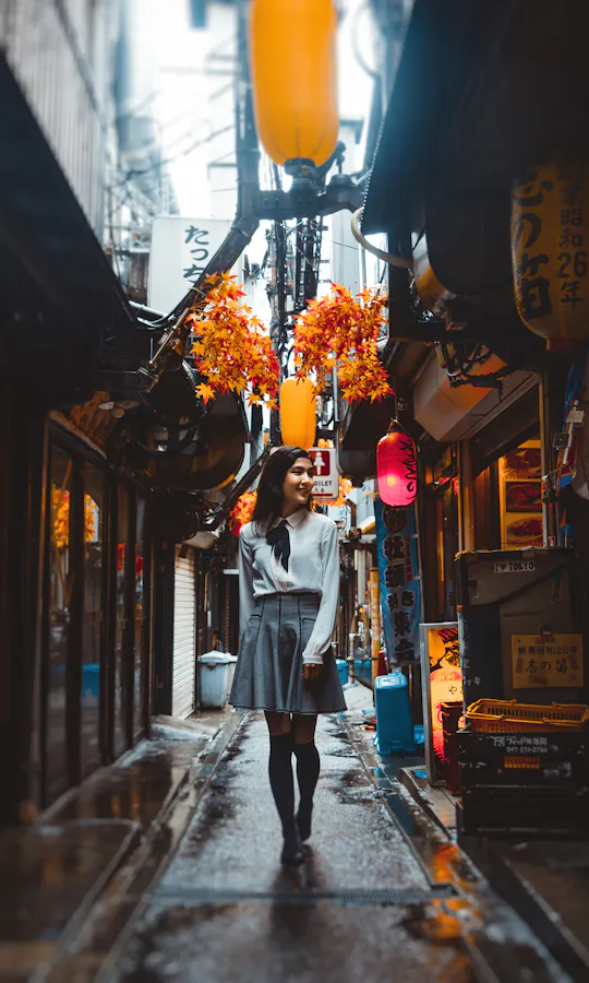 Shinjuku Golden Gai A woman walks down a narrow, lantern-lit alley lined with small shops and restaurants, with colorful signs and hanging decorations, on a rainy day.