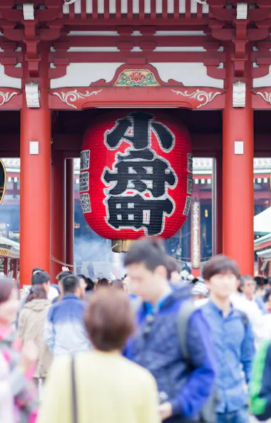 Kaminarimon Gate A large crowd of people walks beneath the iconic red lantern and wooden gate at Senso-ji Temple in Tokyo, Japan, with ornate details and Japanese characters visible above the entrance.