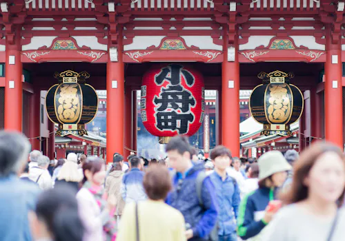 A large crowd of people walks beneath the iconic red lantern and wooden gate at Senso-ji Temple in Tokyo, Japan, with ornate details and Japanese characters visible above the entrance.