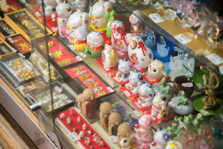 A display case filled with various small figurines, including several maneki-neko (beckoning cat) statues, and assorted trinkets and jewelry arranged neatly on red trays, all with price tags.