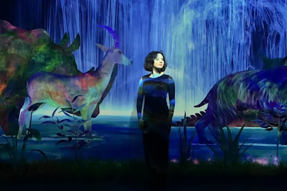 teamLab Planets TOKYO A person stands in front of a digital display of a waterfall with vibrant, colorful images of animals like a goat and a dinosaur. The background is filled with lush plants and a serene environment.