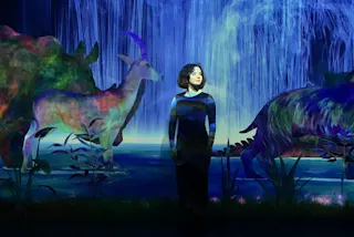A person stands in front of a digital display of a waterfall with vibrant, colorful images of animals like a goat and a dinosaur. The background is filled with lush plants and a serene environment.