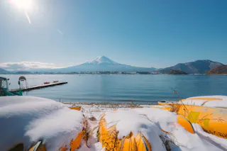 Snow-covered kayaks in the foreground with a calm lake and Mount Fuji in the background. A clear blue sky and distant mountains frame the lake, creating a serene winter scene.