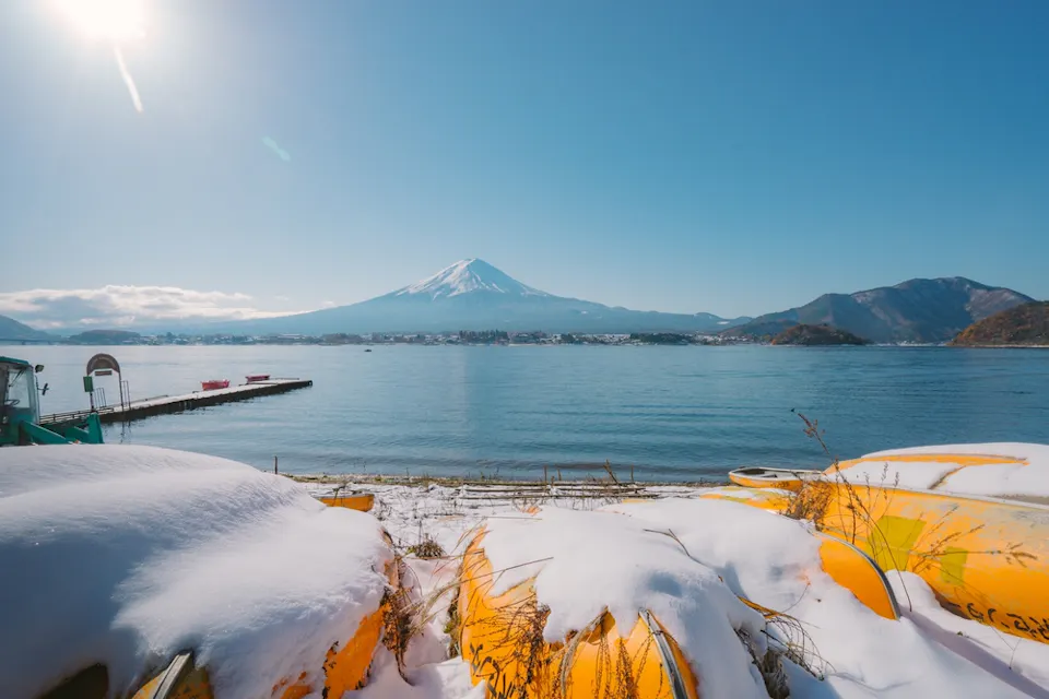 Lake Kawaguchiko Snow-covered kayaks in the foreground with a calm lake and Mount Fuji in the background. A clear blue sky and distant mountains frame the lake, creating a serene winter scene.