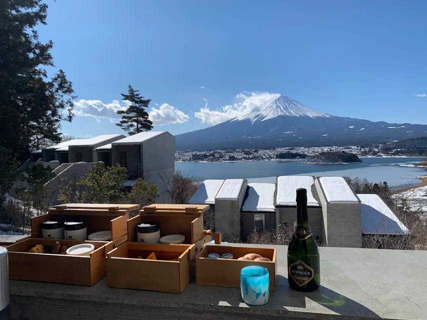 Breakfast boxes and a bottle of champagne on a table overlooking snow-capped Mount Fuji and a lake. Modern buildings are visible in the foreground, surrounded by trees and snow. The sky is clear with a few clouds.