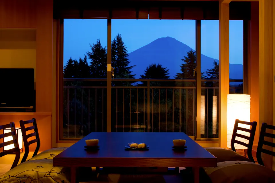 A cozy room with wooden furniture, lit by warm lamps, features a low table with teacups and snacks. Large windows reveal a view of Mount Fuji at dusk, framed by trees.