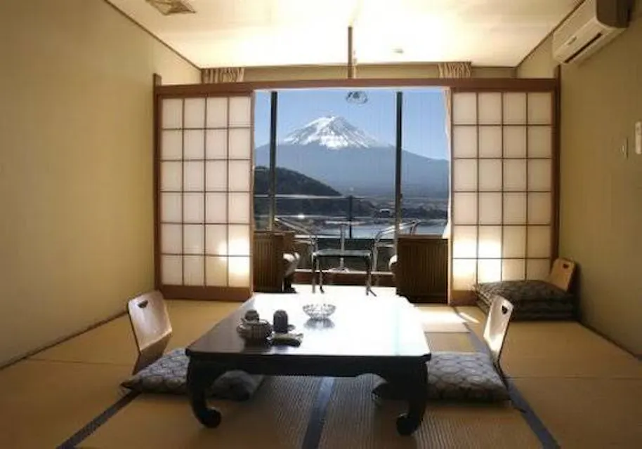 A traditional Japanese room with tatami mats, a low table, and floor cushions. Sliding shoji doors open to a balcony with a view of a snow-capped Mount Fuji under a clear blue sky.