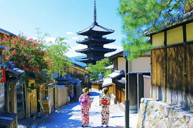 Two people in traditional Japanese clothing walk down a narrow street lined with wooden buildings and trees. A pagoda towers in the distance under a clear blue sky.