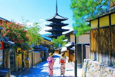Two people in traditional Japanese clothing walk down a narrow street lined with wooden buildings and trees. A pagoda towers in the distance under a clear blue sky.