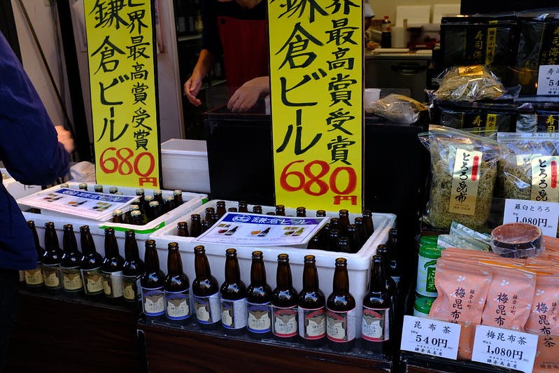 Bottles of beer lined up on a market stall with yellow price signs in Japanese, surrounded by packaged food items, teas, and a person’s hand on the left.