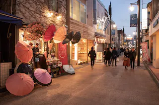 A narrow street at dusk with people walking, illuminated by streetlights. Colorful umbrellas are displayed outside a shop on the left, adding a vibrant touch to the cozy, lively urban scene.