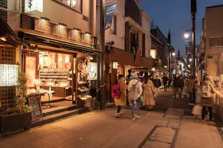 A lively street scene at dusk in Japan shows people walking past warmly lit shops with traditional signs and displays, creating a welcoming and vibrant atmosphere.