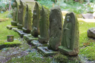A row of small, weathered stone statues with moss growing on them is lined up on a green, mossy ground in a peaceful outdoor setting. Some statues are detailed, while others appear eroded.