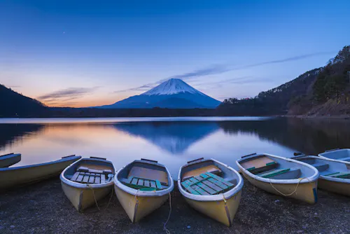 Rowboats rest on the shore of a calm lake at sunset, with Mount Fuji in the background, its snow-capped peak reflected in the still water, surrounded by gentle hills and a clear, blue sky.