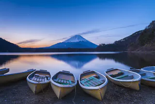 Rowboats rest on the shore of a calm lake at sunset, with Mount Fuji in the background, its snow-capped peak reflected in the still water, surrounded by gentle hills and a clear, blue sky.