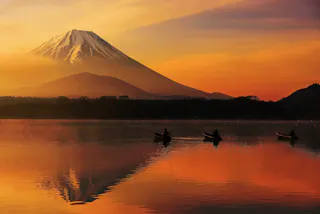 Mount Fuji at sunset, reflected in a calm lake with three people in small boats silhouetted against the orange and golden sky.