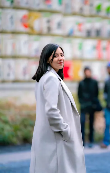 Meiji Shrine A woman in a light-colored coat stands outdoors, smiling at the camera. In the background, three people are blurred, and colorful objects or signs are displayed on a large wall. The scene appears to be in a park or public area.